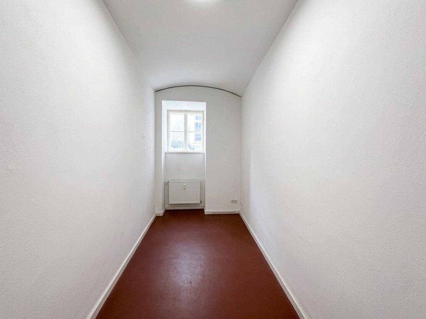 Schmaler, leerer Raum mit dunkelrotem Boden, weißen Wänden und einem Fenster, das Licht spendet.