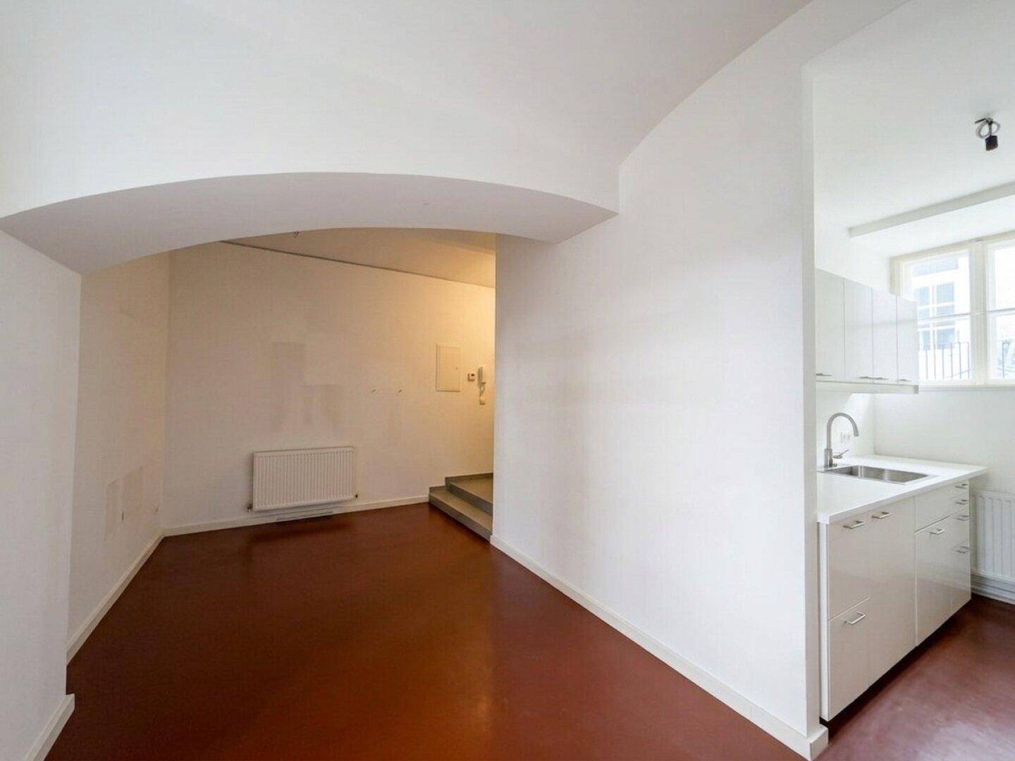 Offener Raum mit dunkelrotem Boden, weiß gestrichenen Wänden und einer kleinen Küchenzeile.