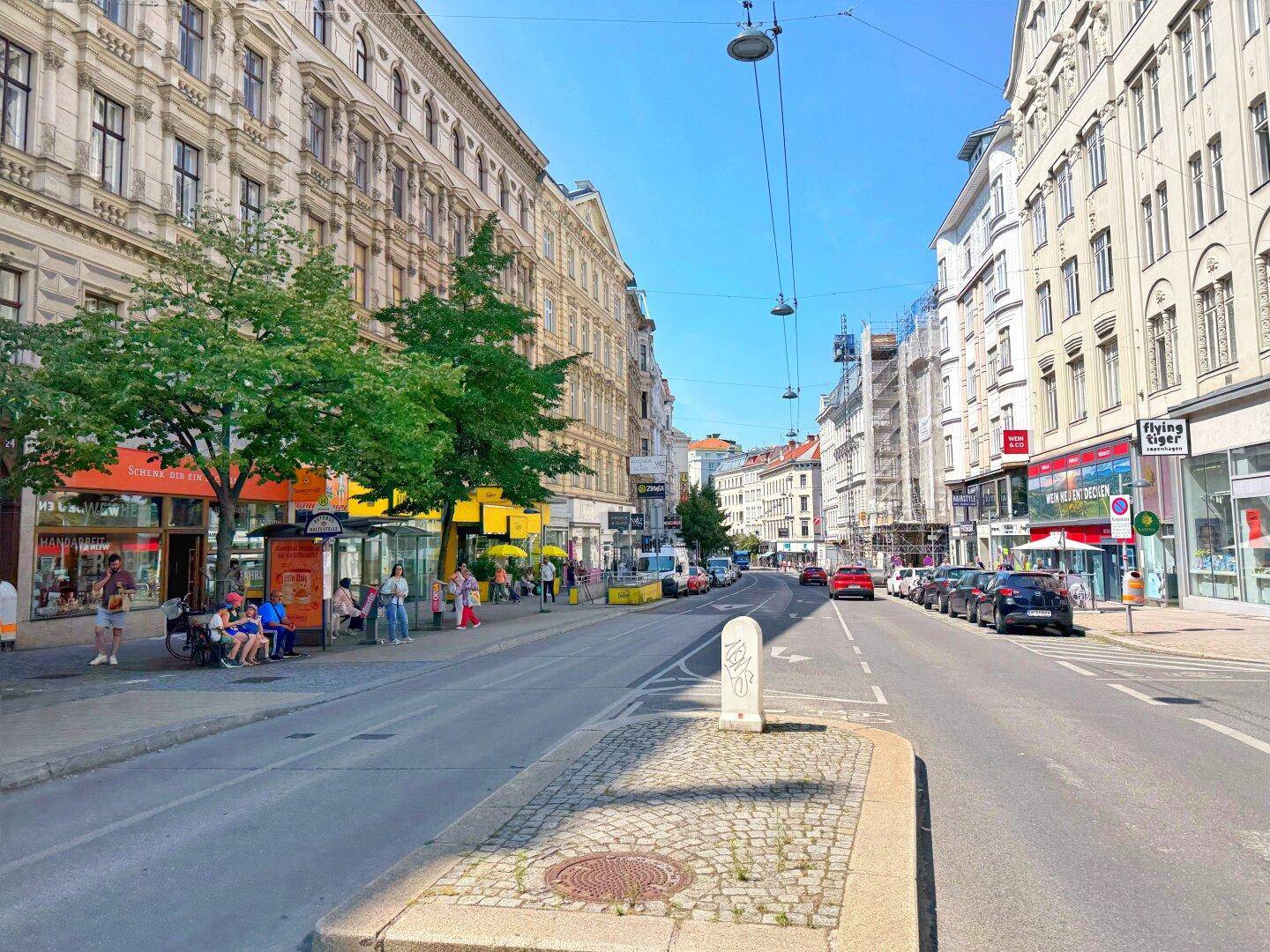 Belebte Stadtstraße mit historischen Gebäuden, parkenden Autos, Bäumen und Fußgängern an einem sonnigen Tag.