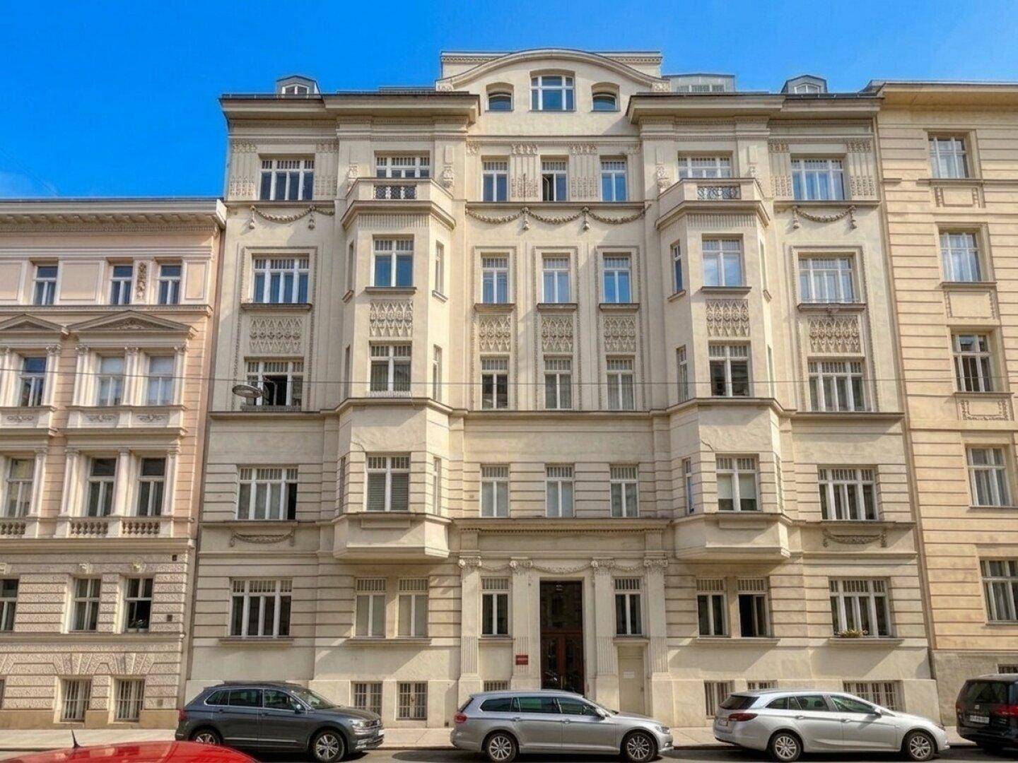 Prächtige Fassade eines mehrstöckigen Gebäudes mit aufwendigen Details und zahlreichen Fenstern, davor parken Autos.