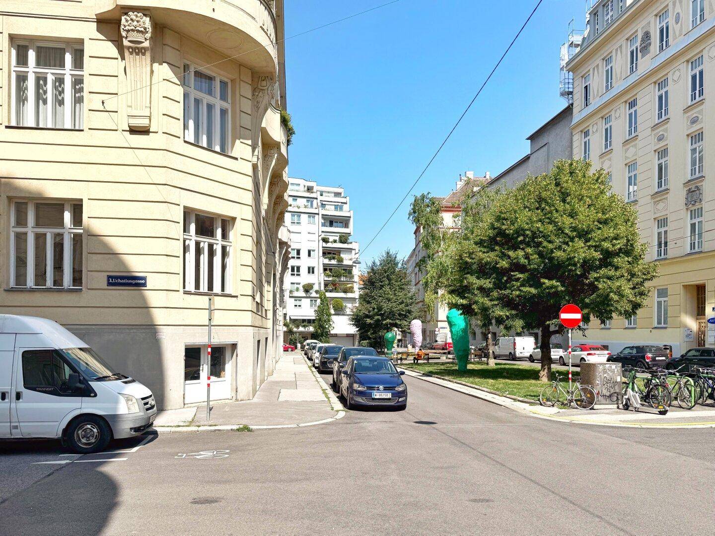 Straßenecke mit Gebäuden, Bäumen und geparkten Autos unter einem klaren blauen Himmel an einem sonnigen Tag.