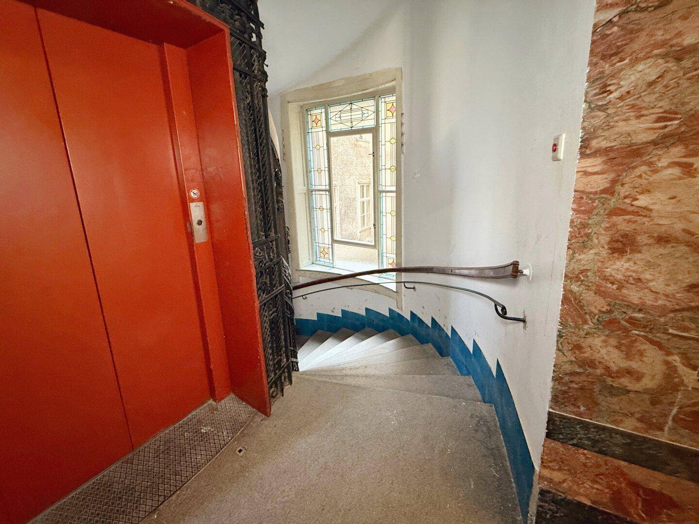 Historisches Treppenhaus mit einem roten Aufzug, einer Wendeltreppe und einem Buntglasfenster, das natürliches Licht hereinlässt.