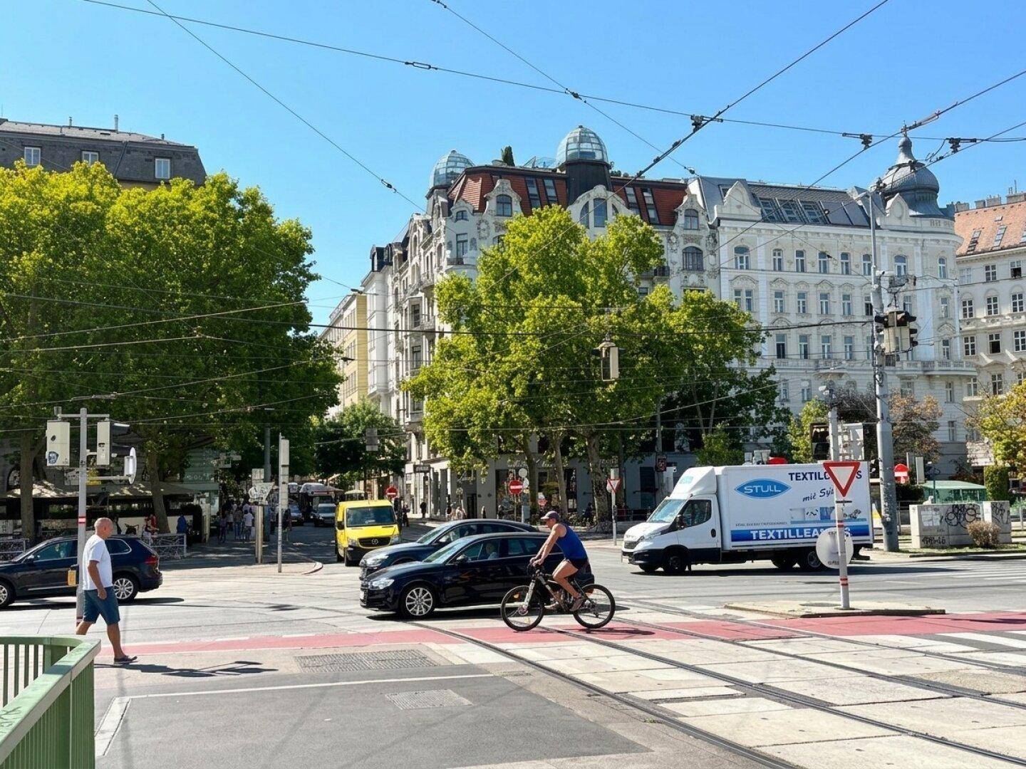 Belebte Straßenszene an einem sonnigen Tag mit Verkehr, Fußgängern und historischen Gebäuden.