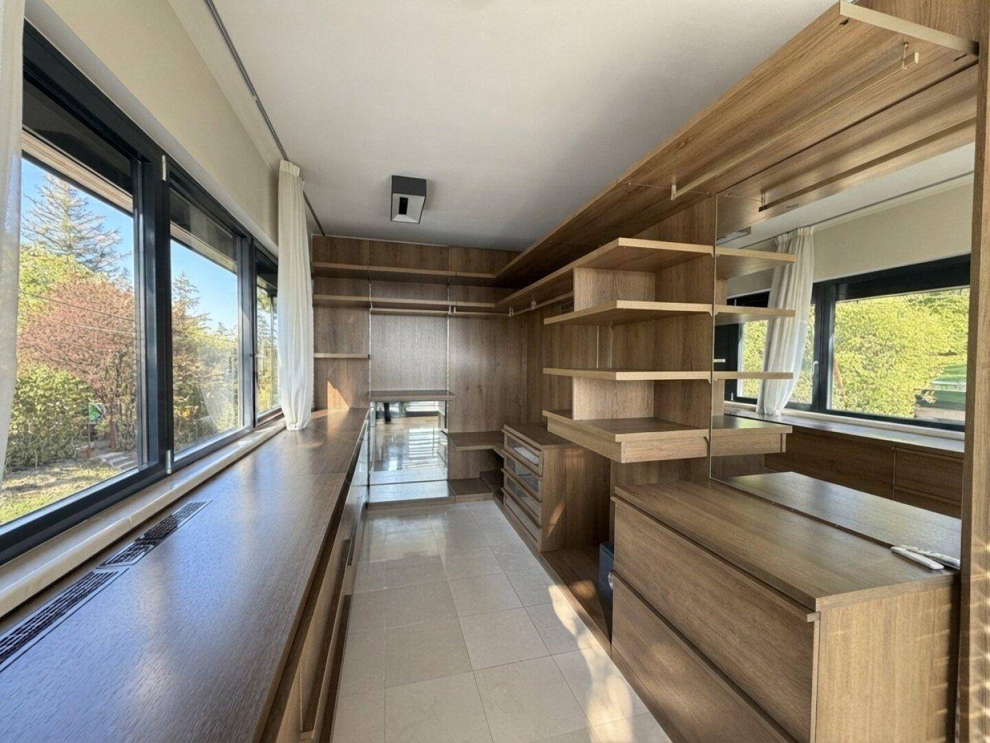 Heller begehbarer Kleiderschrank mit viel Stauraum und großen Fenstern mit Blick ins Grüne.