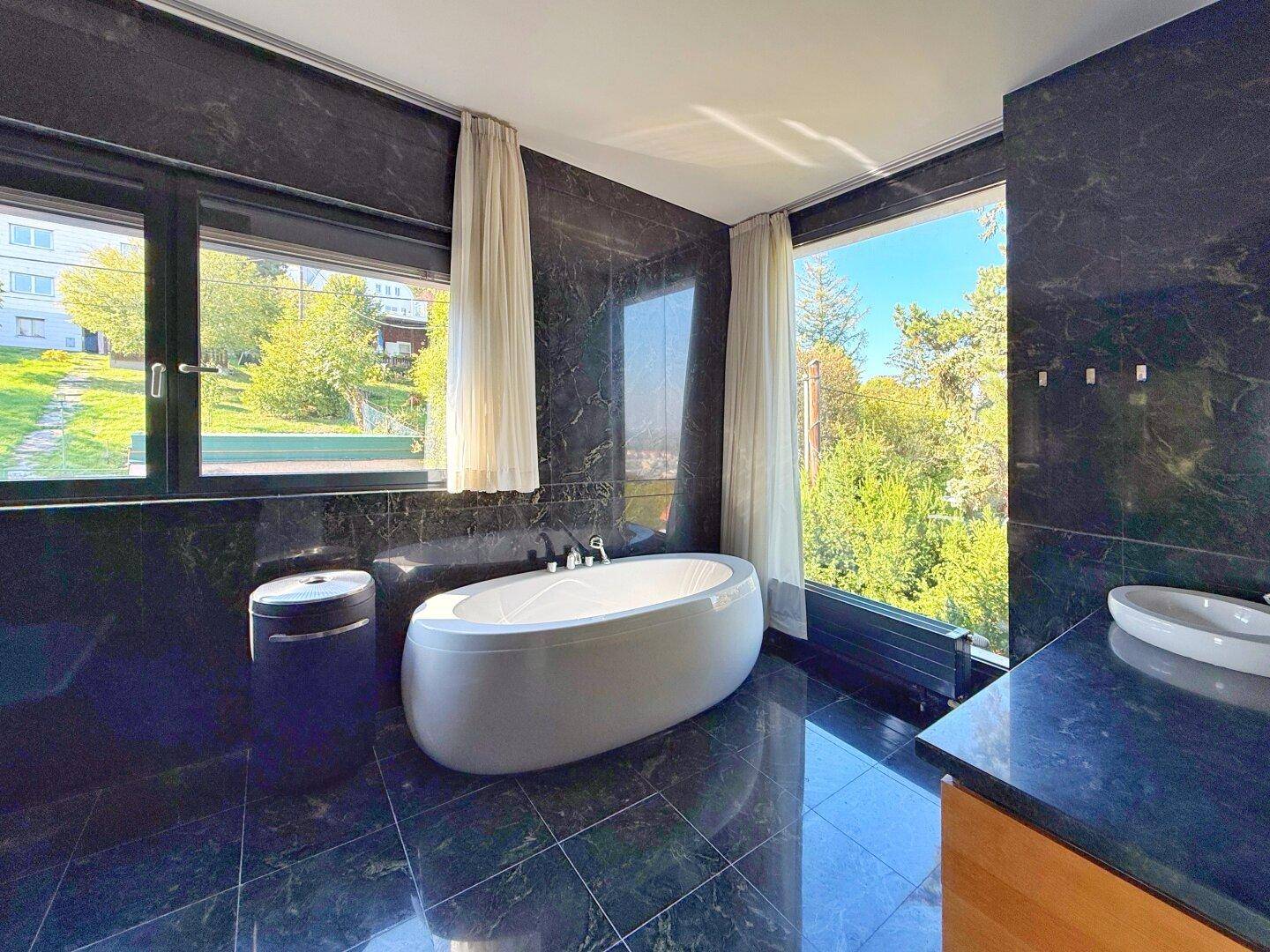 Luxuriöses Badezimmer mit freistehender Badewanne, dunklem Marmor und großen Fenstern mit Grünblick.