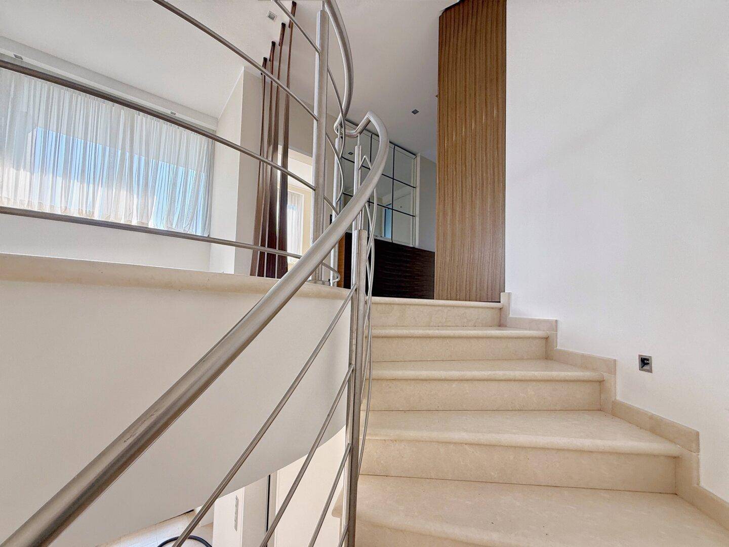 Elegante geschwungene Treppe mit Edelstahlgeländer und hellem Steinboden, die die Etagen verbindet.