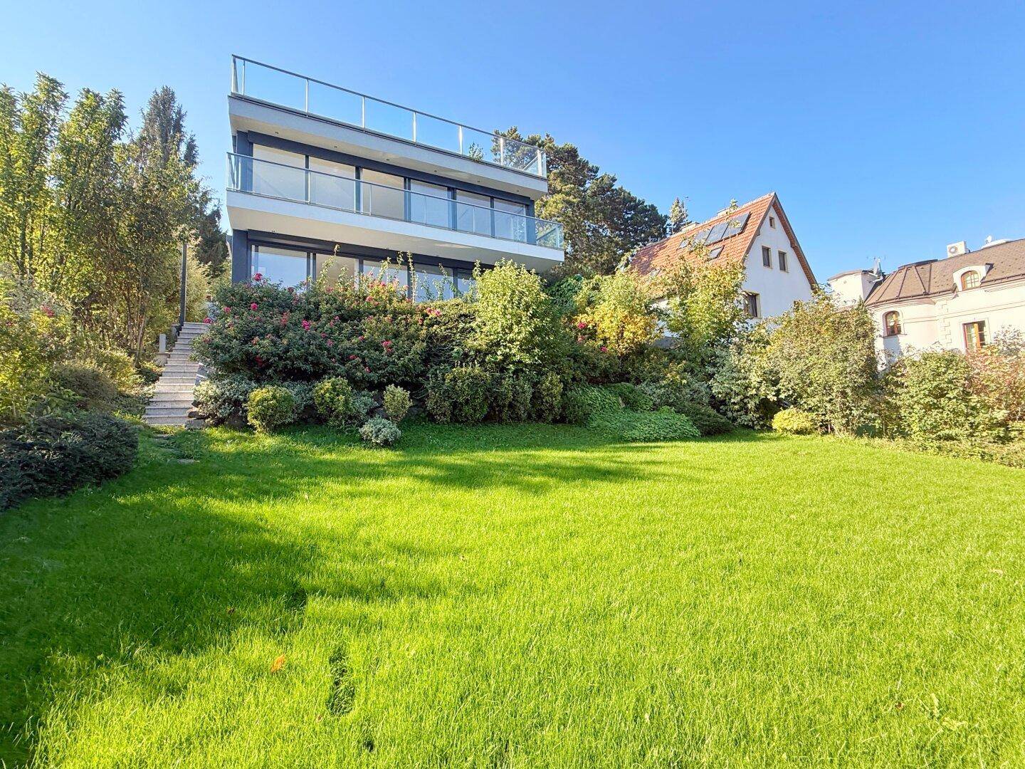 Weitläufiger Rasenbereich vor der modernen Villa mit üppiger Bepflanzung und altem Baumbestand.