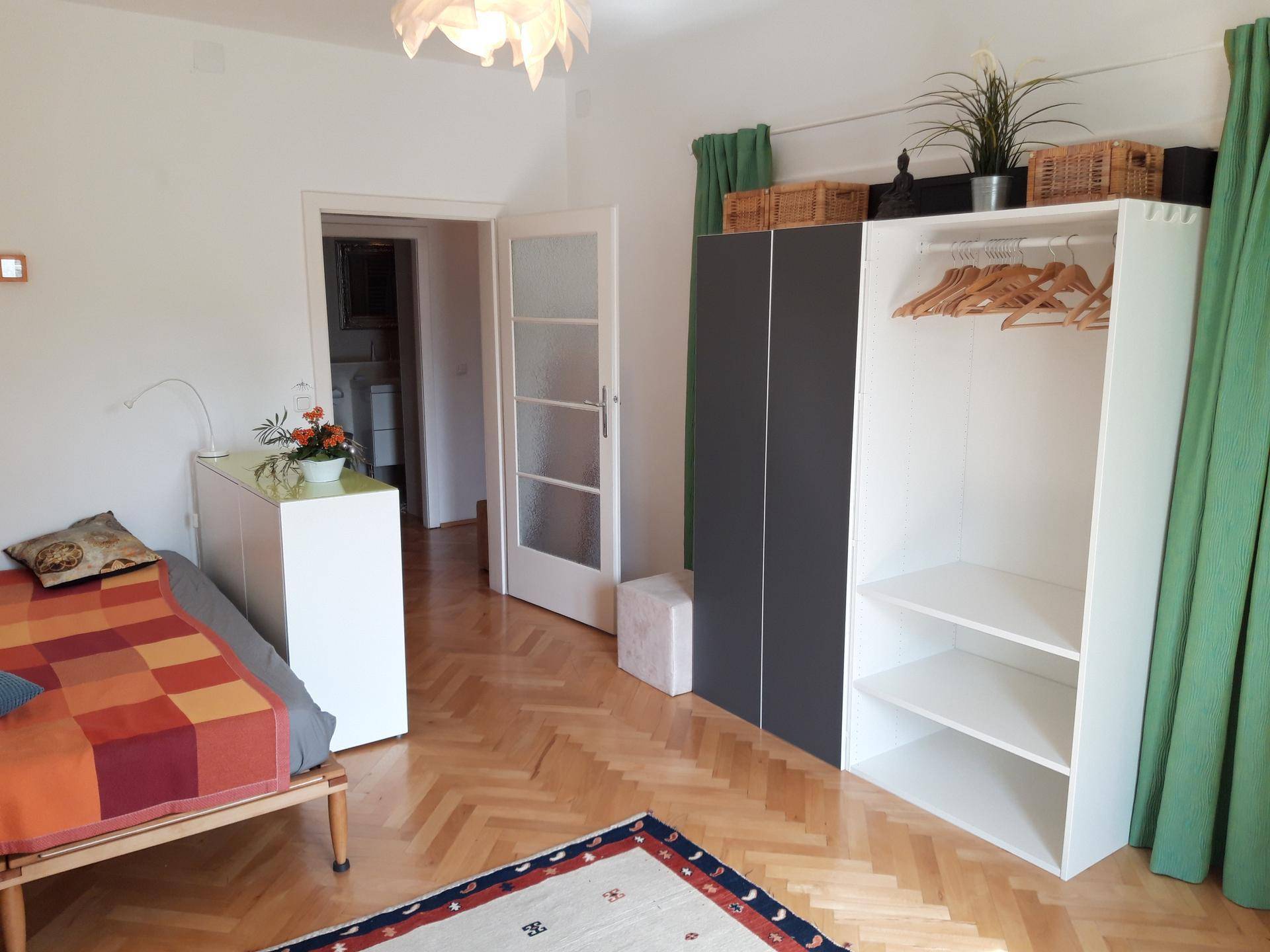 Schlafzimmer mit offenem Kleiderschrank, Holzboden und Zugang zu einem weiteren Raum.