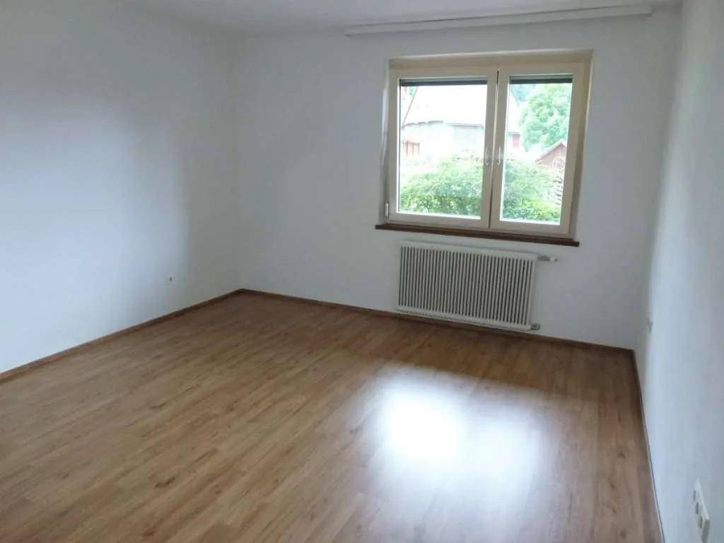 Geräumiges Zimmer mit dunklerem Laminatboden und Fenster mit Blick ins Grüne.
