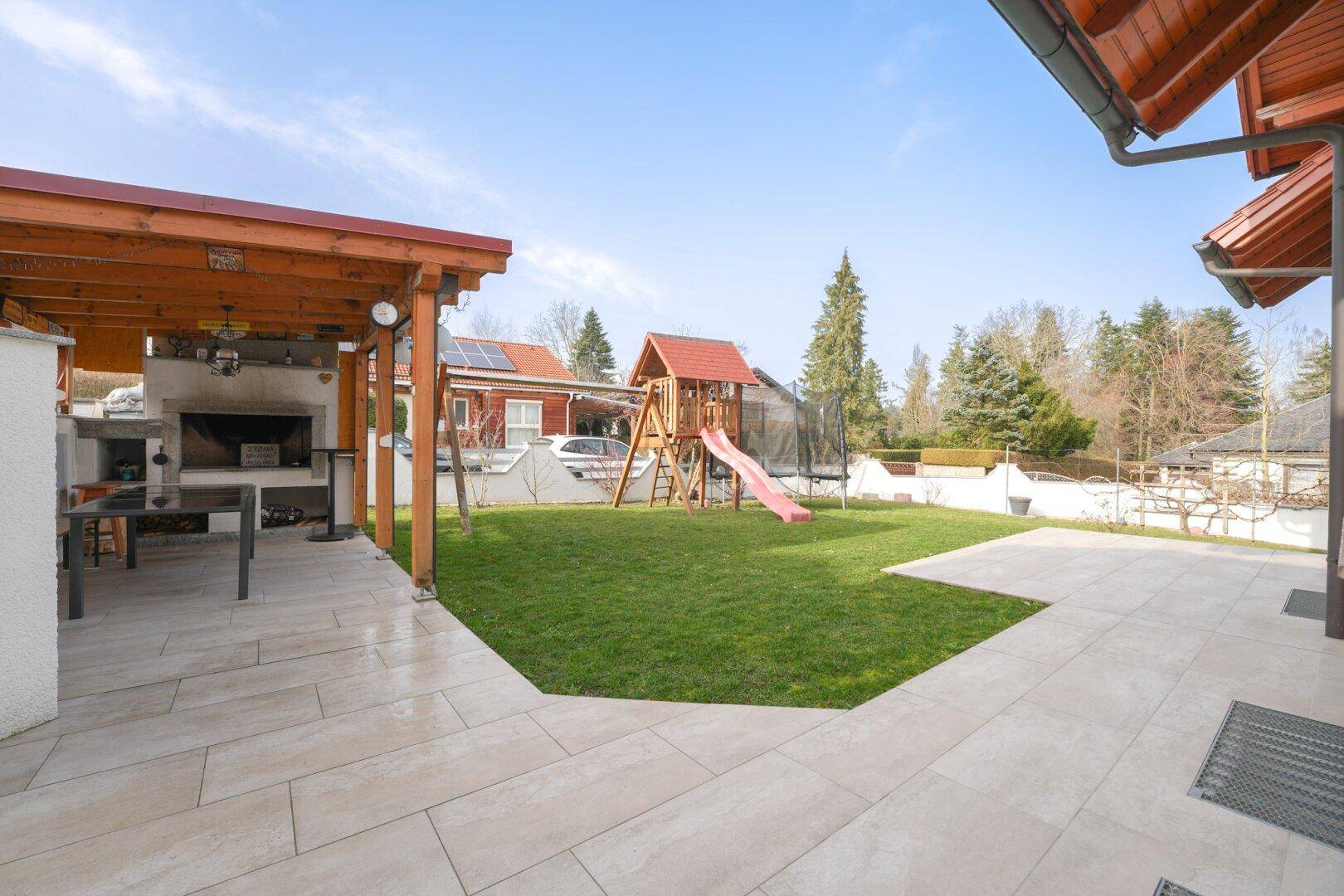 Gartenbereich mit überdachter Außenküche, Grillplatz und Spielgeräten auf dem Rasen.