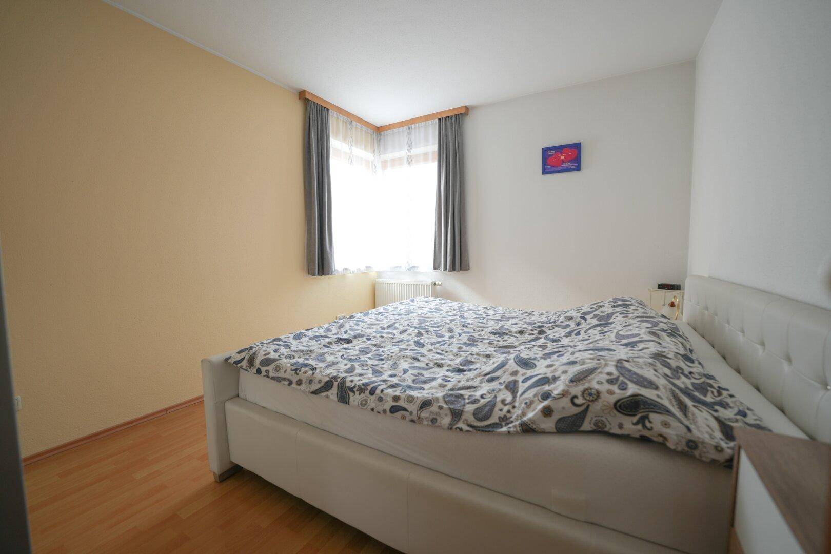 Schlafzimmer mit Doppelbett, Parkettboden und hellem Fenster für viel Tageslicht.