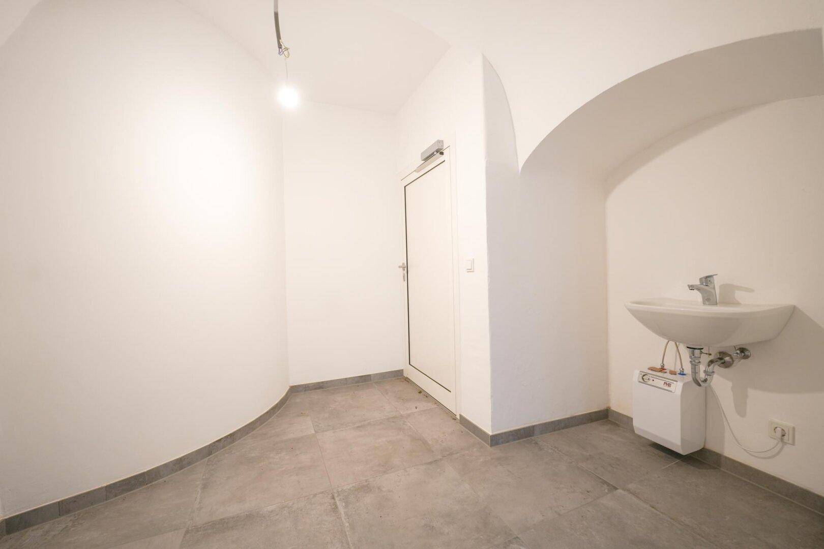 Kleiner Raum mit Waschbecken, weißen Wänden und Fliesenboden, ideal als Gäste-WC oder Abstellraum.