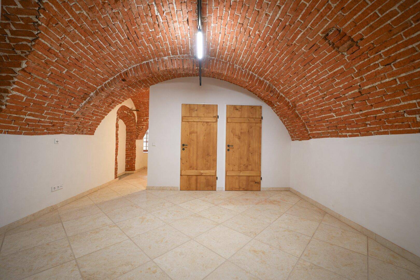 Raum mit beeindruckendem Ziegelgewölbe an der Decke, hellem Fliesenboden und zwei rustikalen Holztüren.