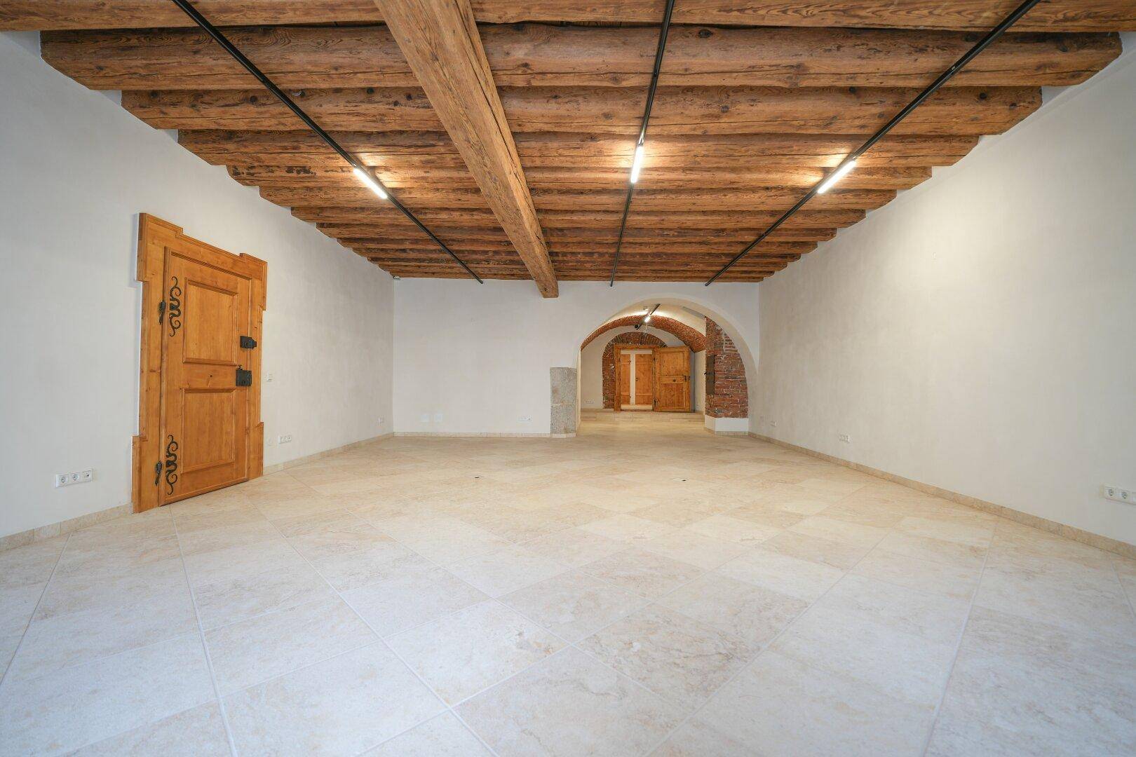 Geräumiger leerer Raum mit sichtbaren Holzbalken an der Decke, hellem Fliesenboden und einem gemauerten Bogen.