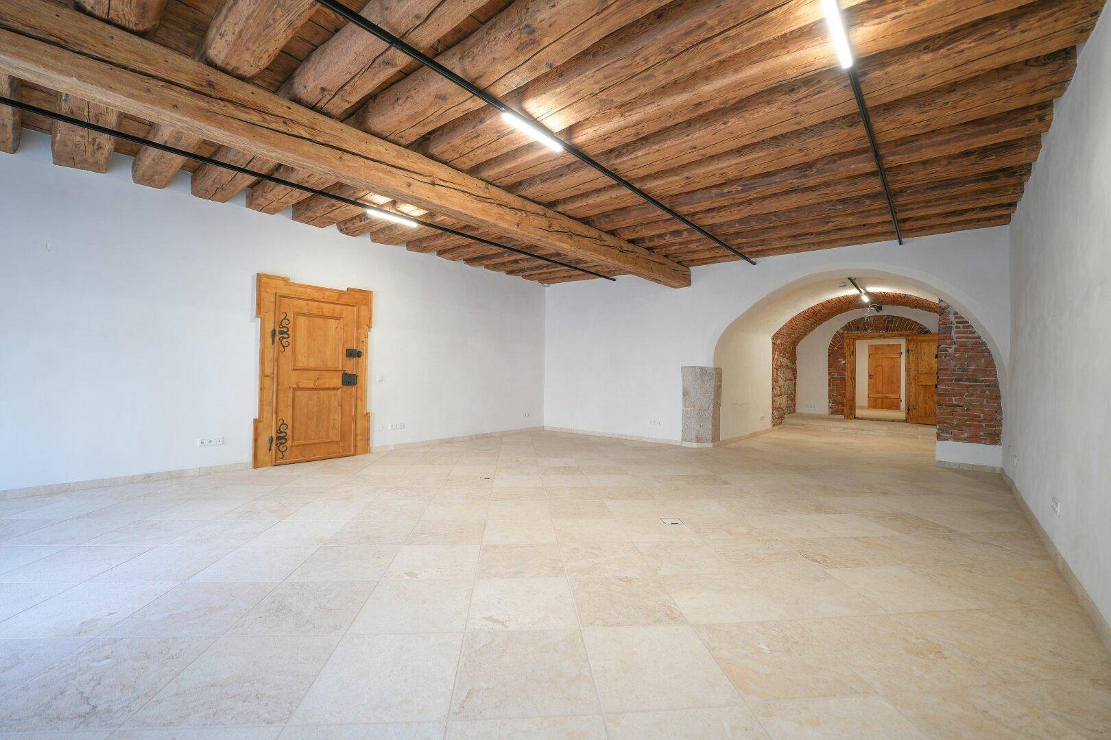 Geräumiger leerer Raum mit sichtbaren Holzbalken an der Decke, hellem Fliesenboden und einem gemauerten Bogen.