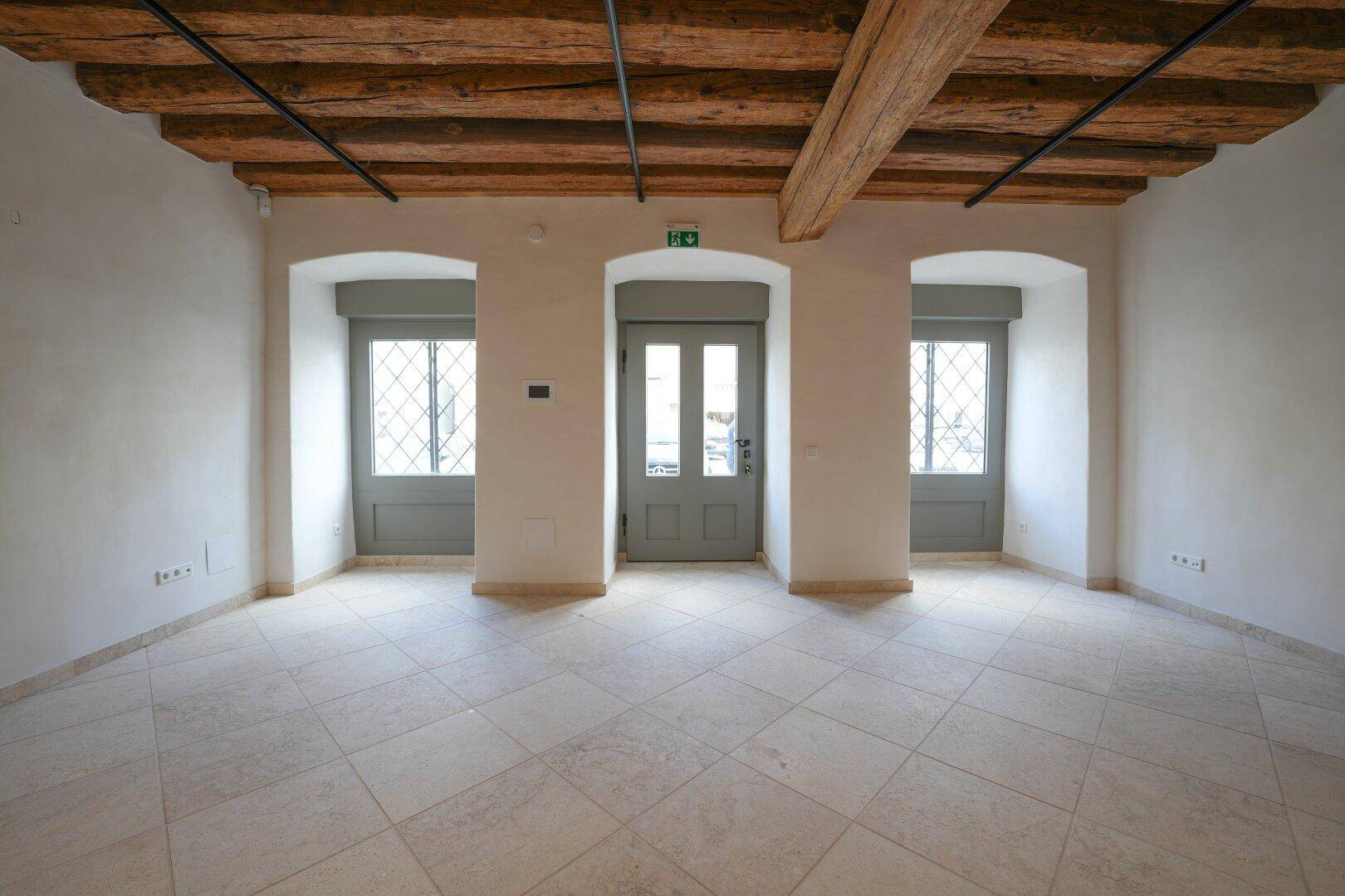 Heller Innenraum mit Holzbalkendecke, Fliesenboden und drei gewölbten Fenstertüren mit Rollos.