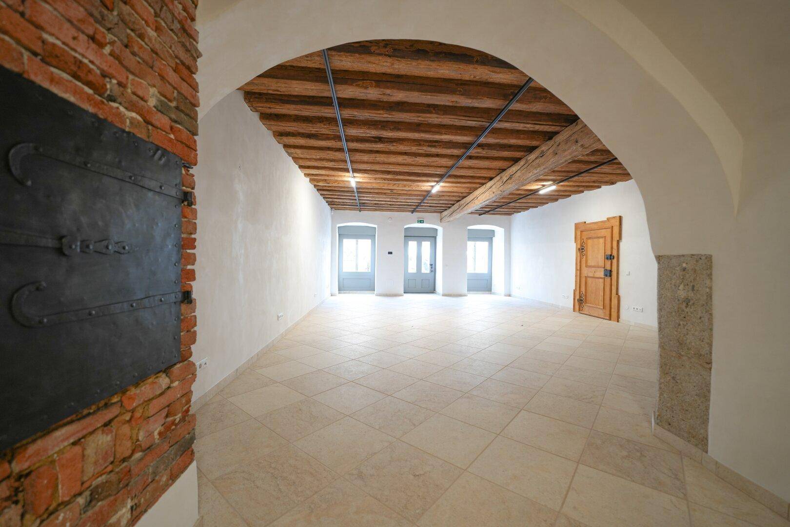 Großer, heller Raum mit sichtbaren Holzbalken, Fliesenboden und mehreren Fenstertüren, mit Ziegelbogen links.