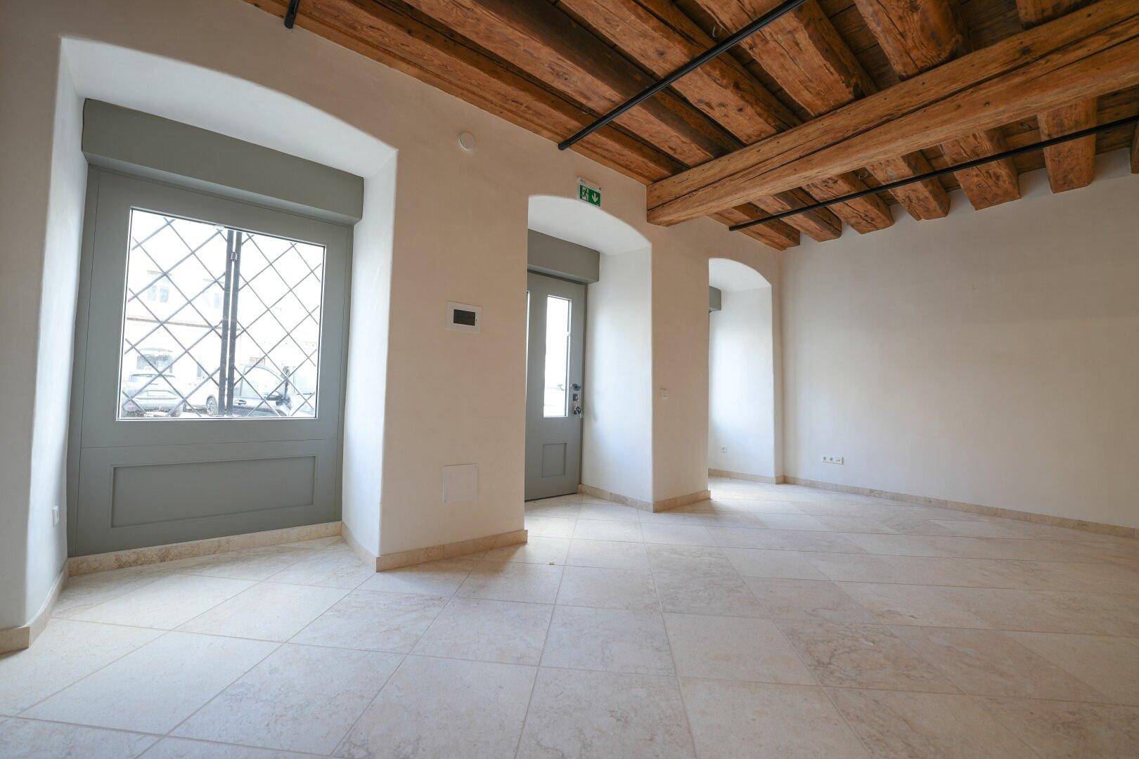 Heller Innenraum mit Holzbalkendecke, Fliesenboden und drei gewölbten Öffnungen, zwei davon mit Fenstertüren.