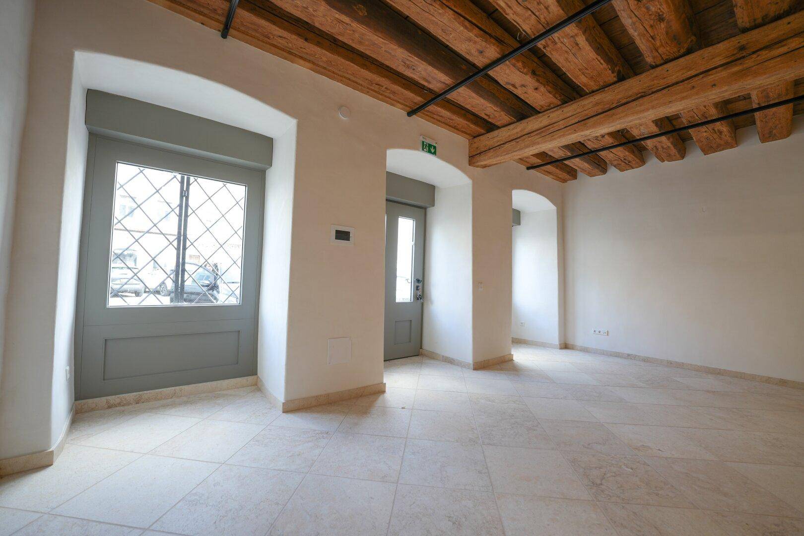 Heller Innenraum mit Holzbalkendecke, Fliesenboden und drei gewölbten Öffnungen, zwei davon mit Fenstertüren und Rollos.