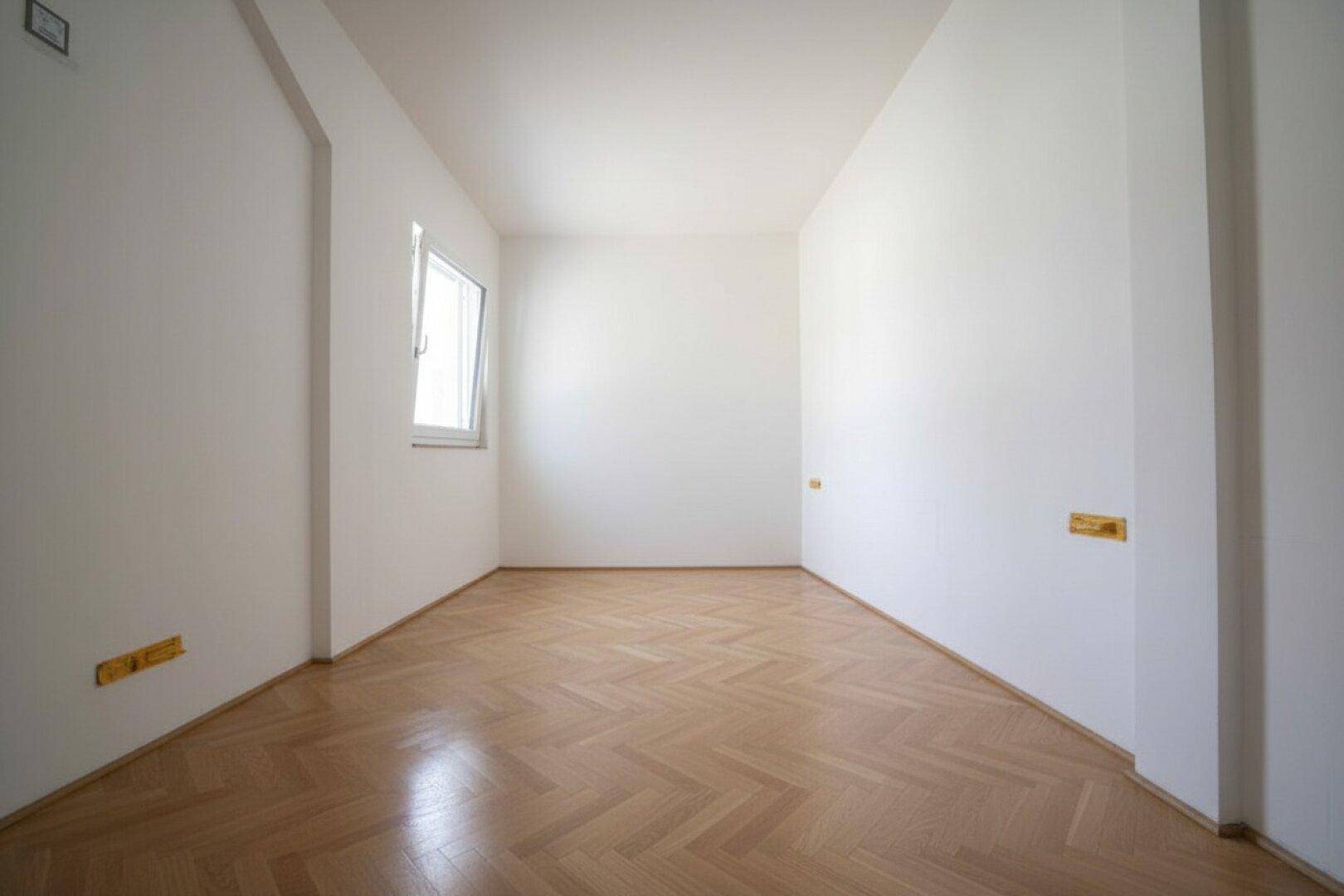 Helles, leeres Zimmer mit Fischgrätparkett und einem Fenster, ideal als Schlaf- oder Arbeitszimmer.