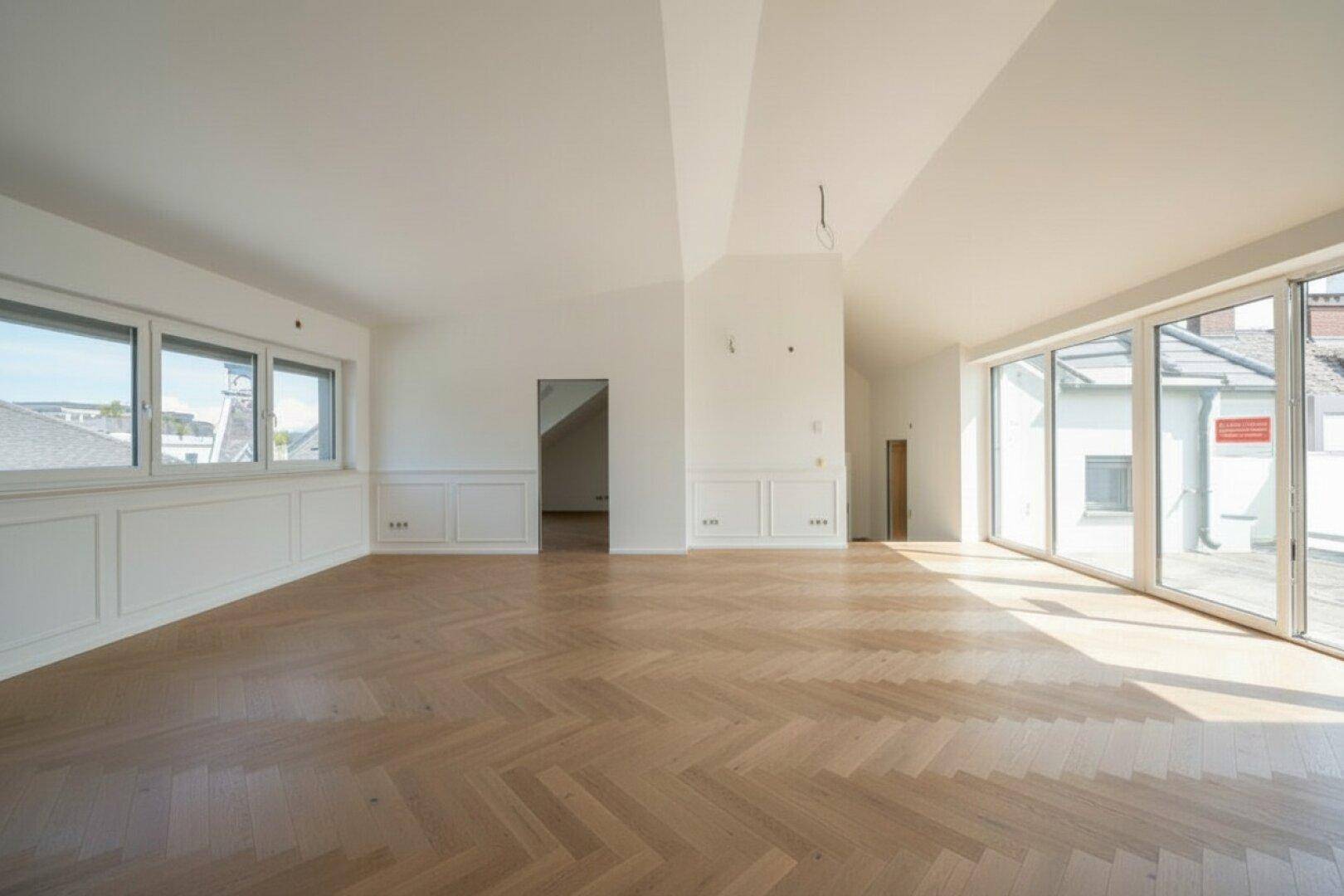 Heller, leerer Wohnraum mit schrägen Decken, Fischgrätparkett und großen Fensterfronten.