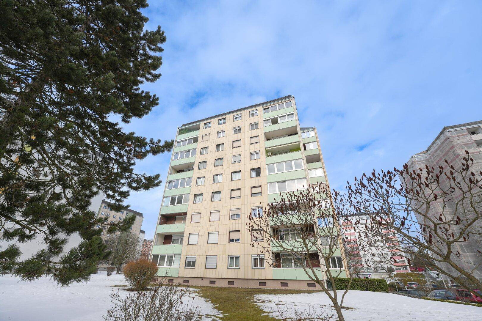 Mehrstöckiges Wohngebäude mit Balkonen und Fenstern, umgeben von Bäumen und Schnee.