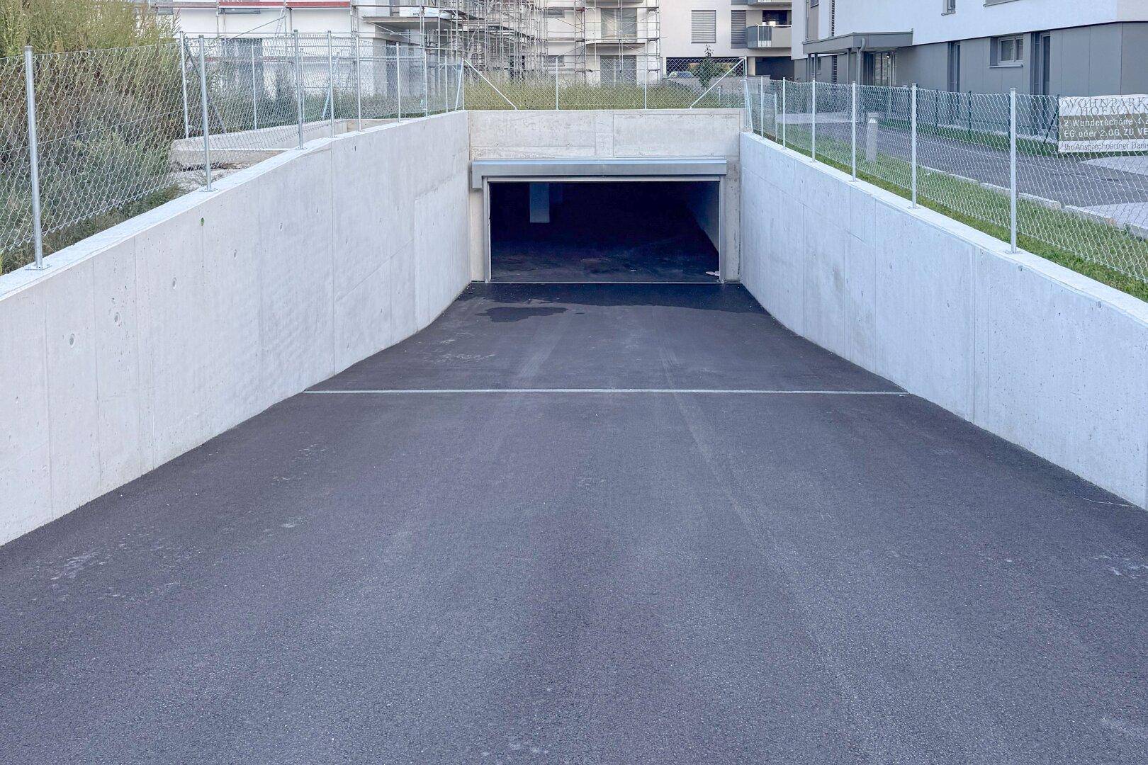 Einfahrt zur Tiefgarage mit Betonwänden und Asphaltrampe, umgeben von einem Zaun.
