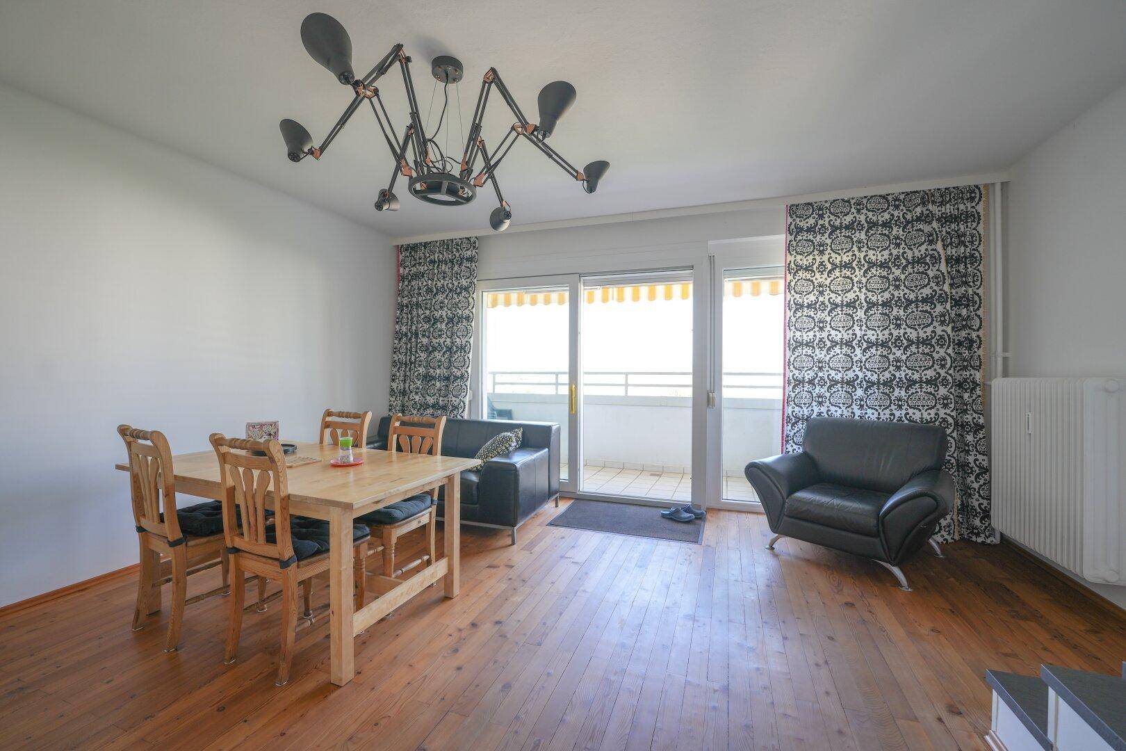 Heller Wohn- und Essbereich mit Holzboden, modernem Kronleuchter und Zugang zum Balkon.