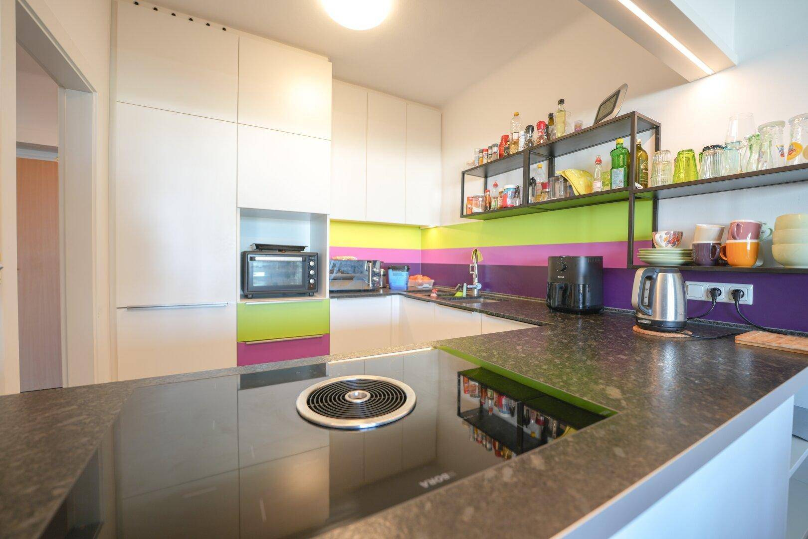 Moderne Küche mit dunkler Arbeitsplatte, Induktionskochfeld und farbigen Akzenten an der Wand.