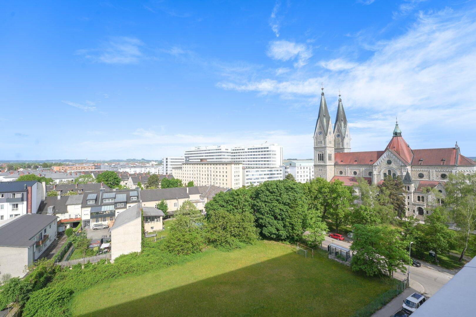 Weitläufiger Blick über eine grüne Wiese, Wohngebäude und eine markante Kirche in der Ferne.