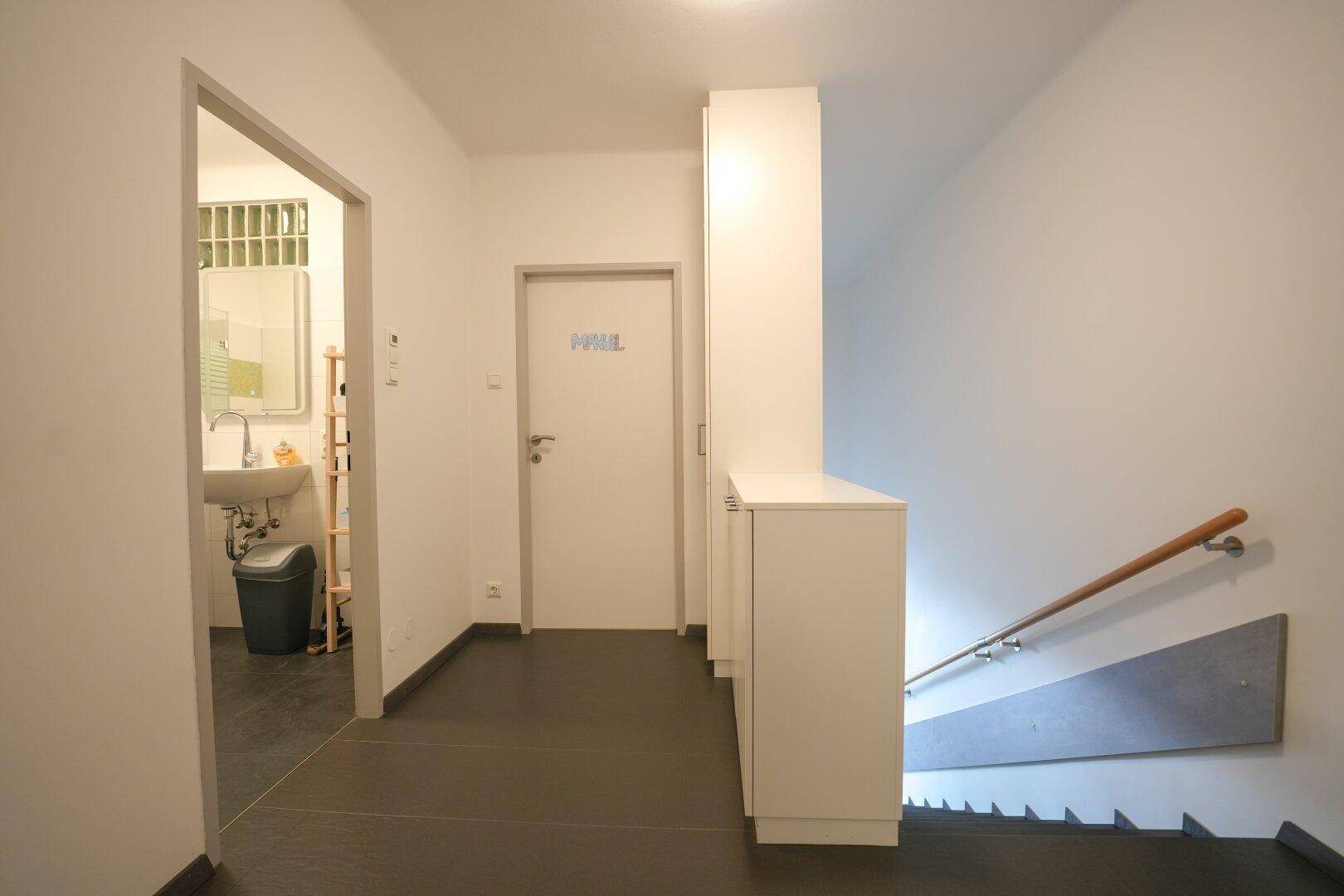 Flur mit dunklem Fliesenboden, Treppenaufgang und Zugang zum Badezimmer.