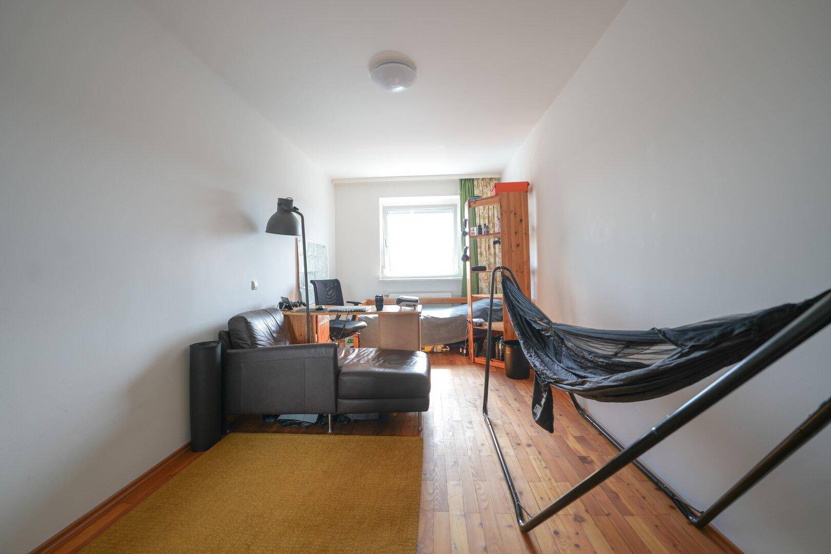 Vielseitiger Raum mit Holzboden, Sofa, Schreibtischbereich und einem Fenster für natürliches Licht.