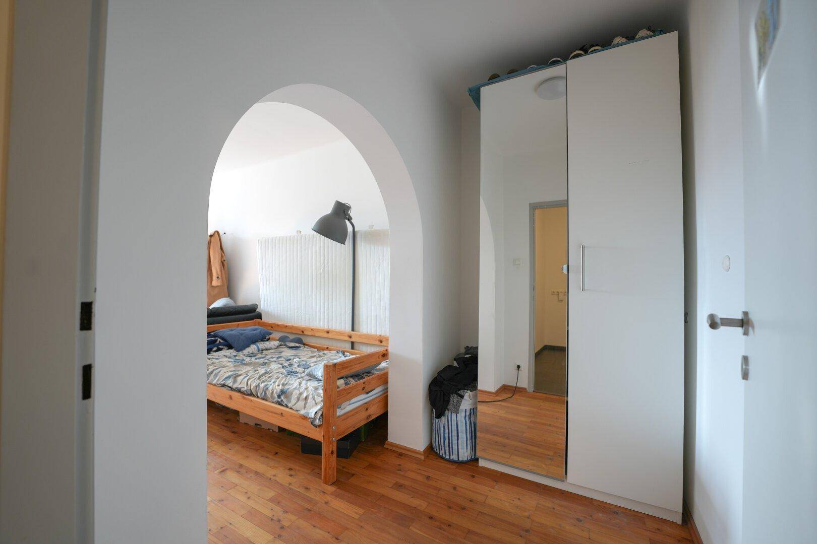 Schlafzimmer mit Holzboden, Einzelbett und einem großen Spiegelschrank, verbunden durch einen Torbogen.