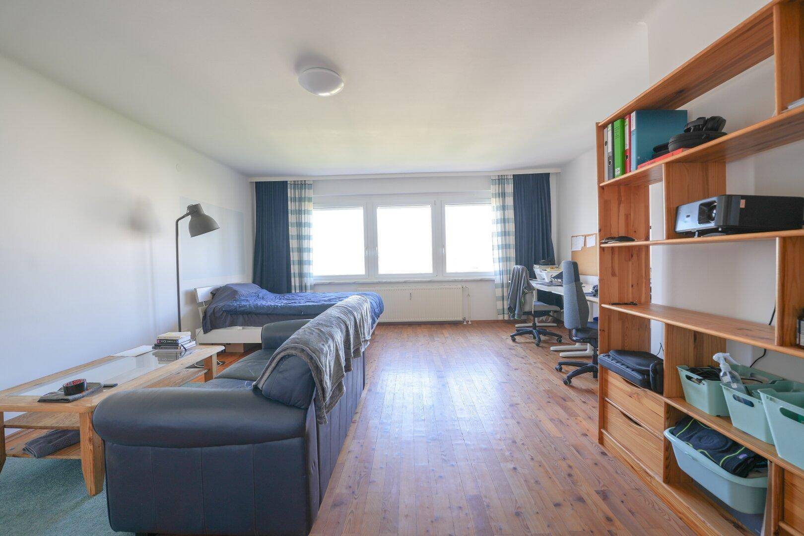 Langer Schlaf- und Wohnbereich mit Holzboden, großem Fenster und funktionaler Einrichtung.