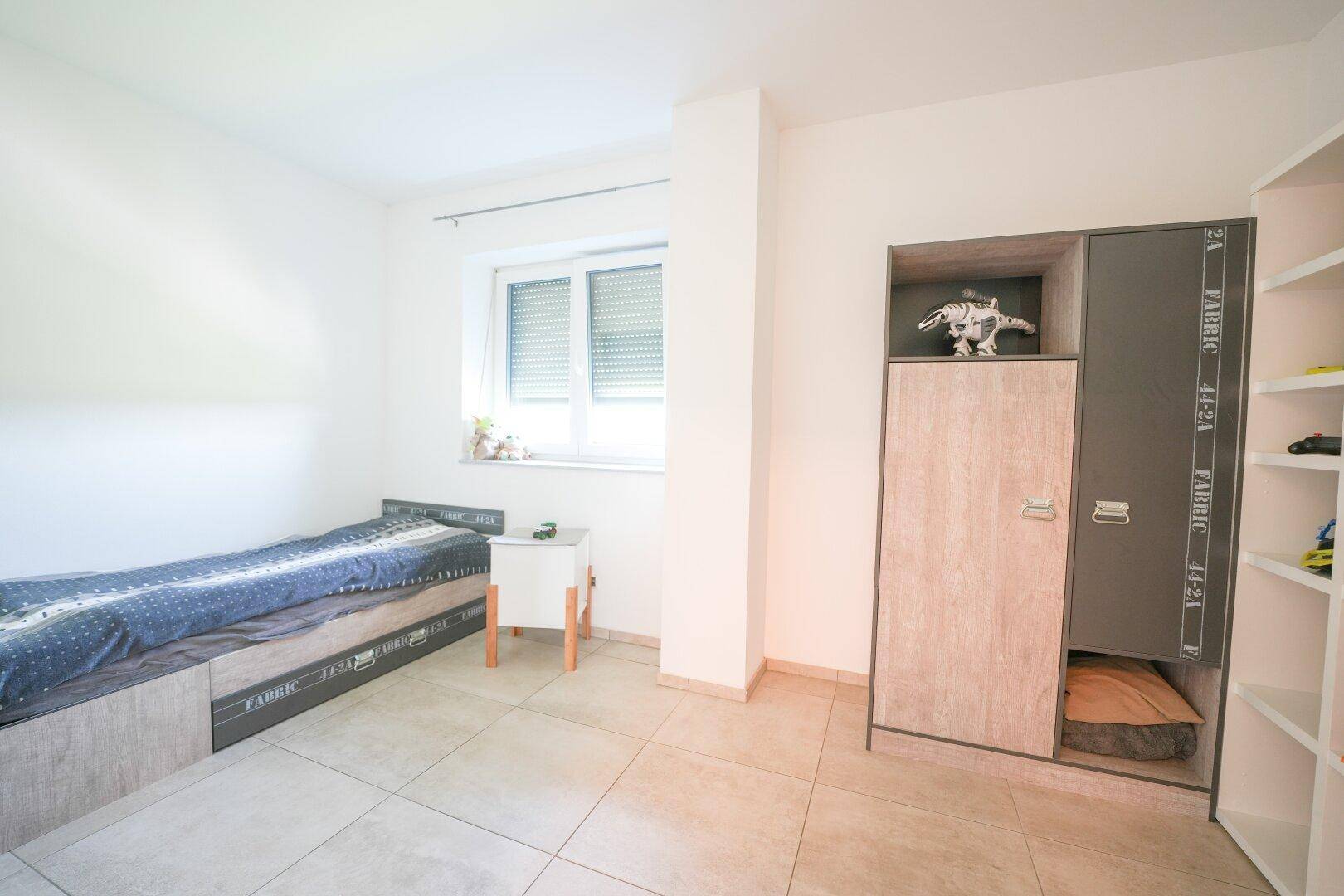 Helles Kinderzimmer mit Einzelbett, Kleiderschrank und Fenster mit Rollladen.