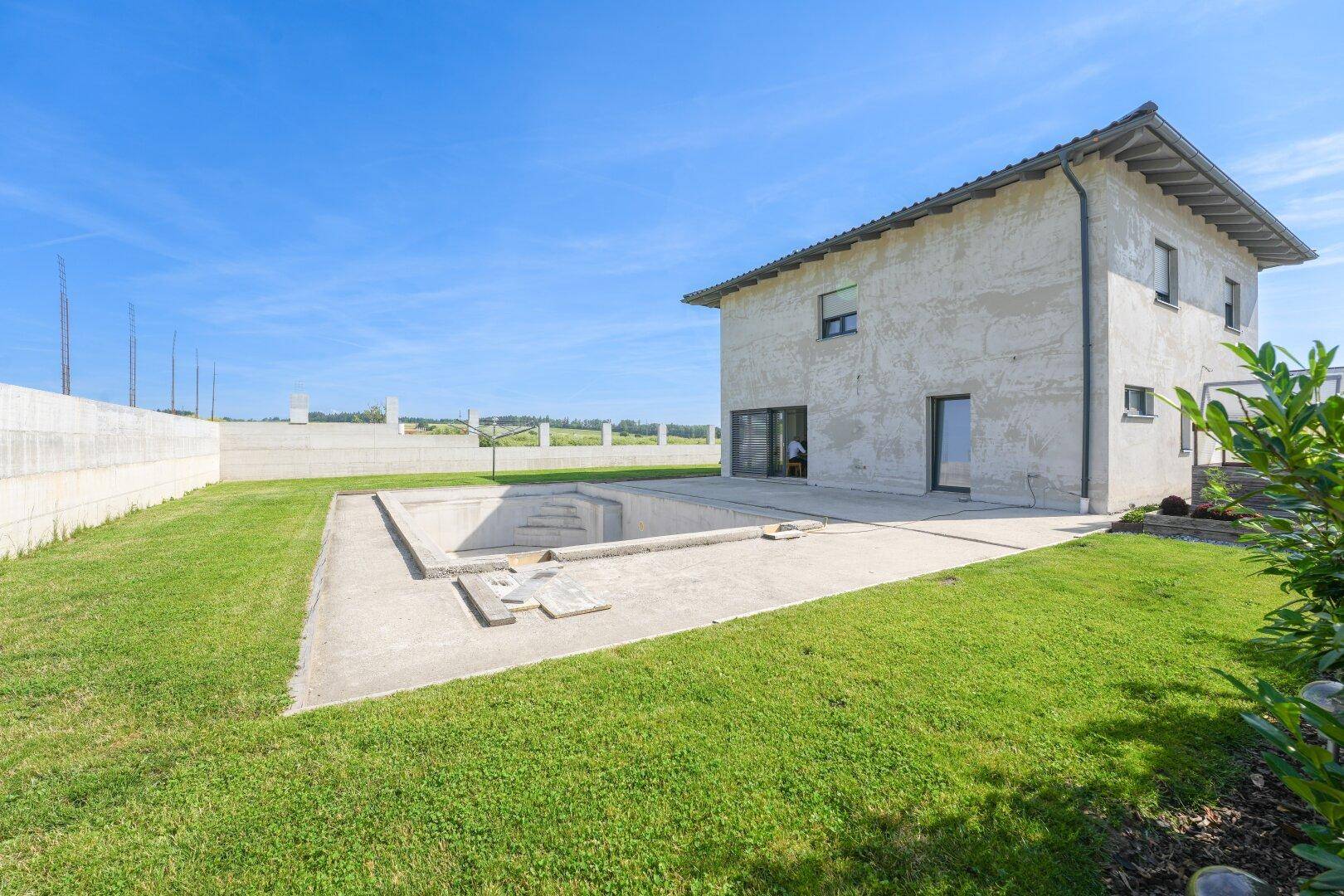 Seitenansicht des Hauses mit weitläufigem Rasen, einer Terrasse und einem unfertigen Poolbereich.