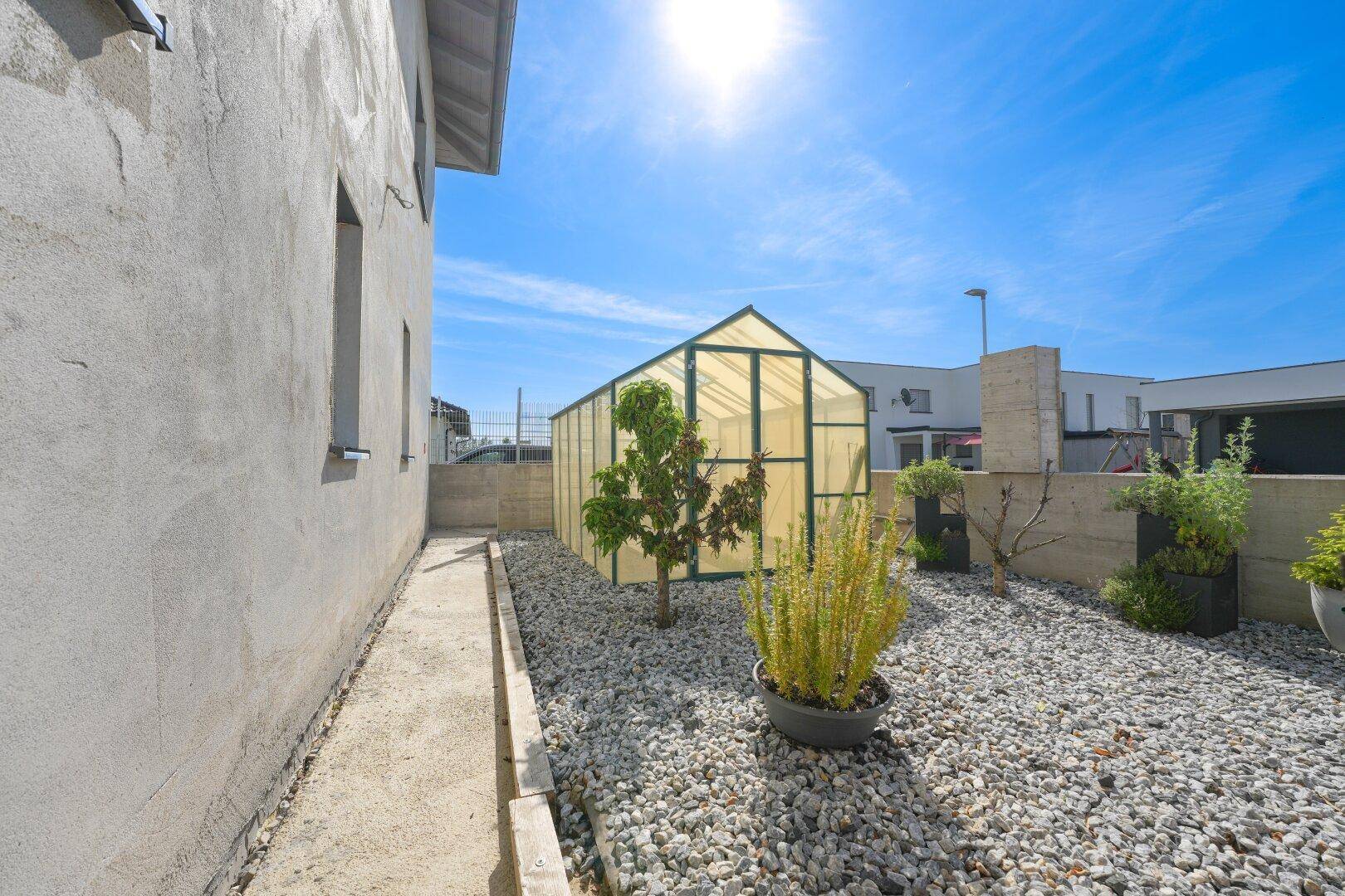Seitlicher Gartenbereich mit Kiesbeet, verschiedenen Pflanzen und einem kleinen Gewächshaus.