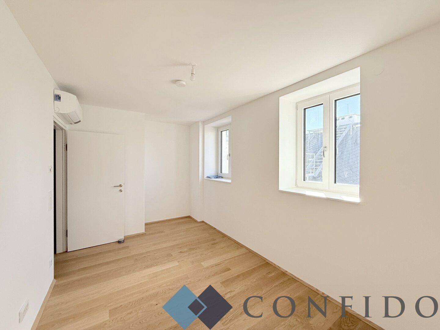 Freundliches Zimmer mit Parkettboden, Klimaanlage und zwei Fenstern, geeignet als Schlaf- oder Gästezimmer.