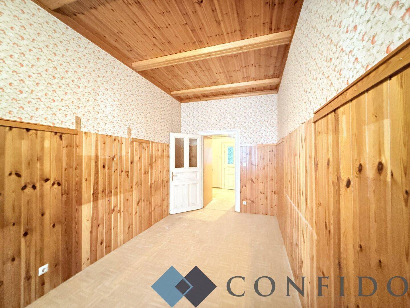 Helles Zimmer mit Holzvertäfelung und gemusterter Tapete, das auf eine Renovierung wartet.