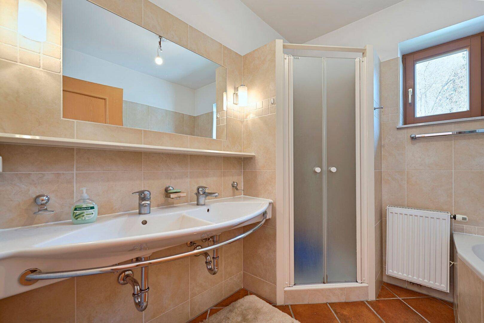Badezimmer mit beigefarbenen Fliesen, Doppelwaschbecken, Duschkabine und einem Fenster.