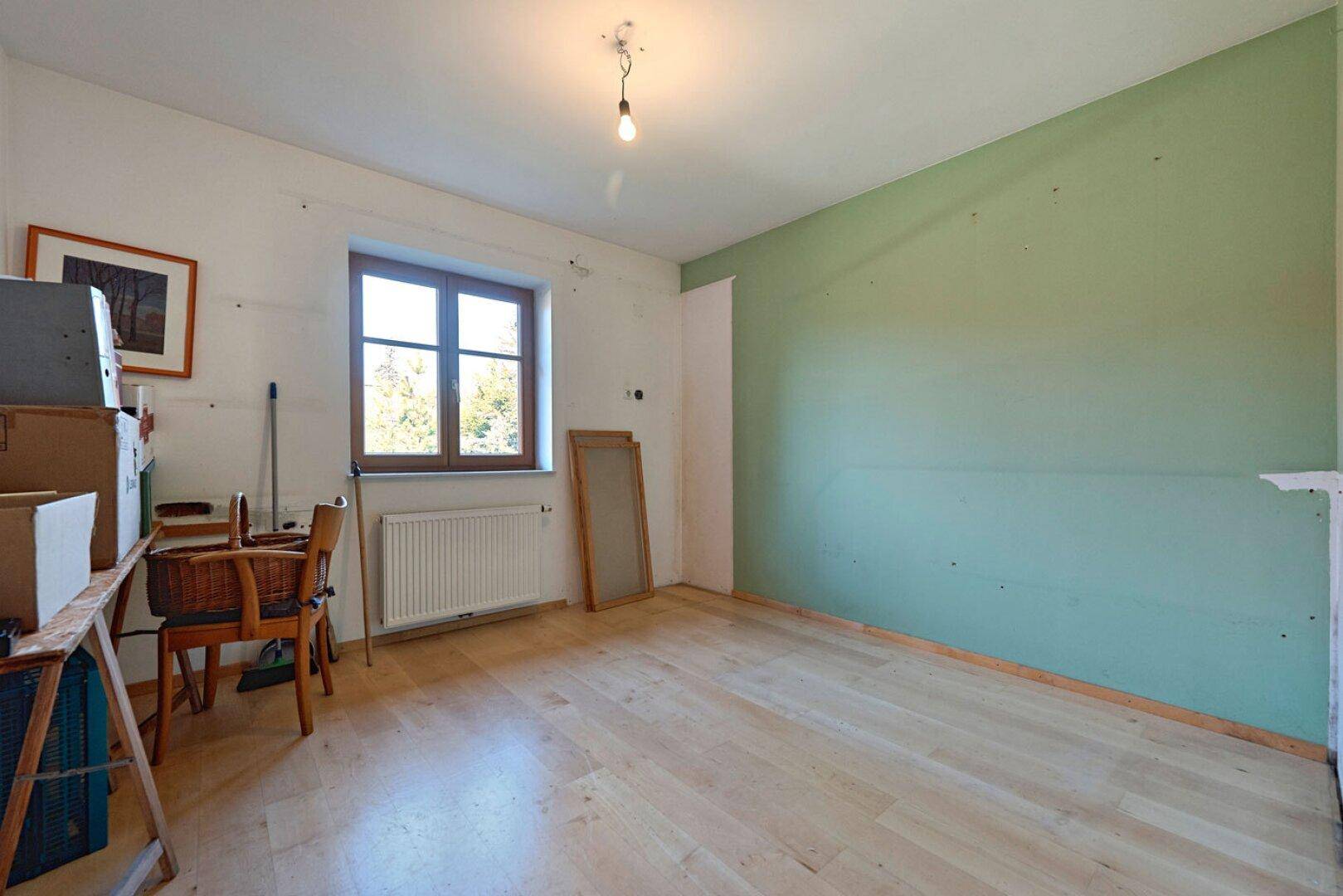 Raum mit hellem Holzboden, grüner Akzentwand und Fenster, derzeit als Abstellraum genutzt.