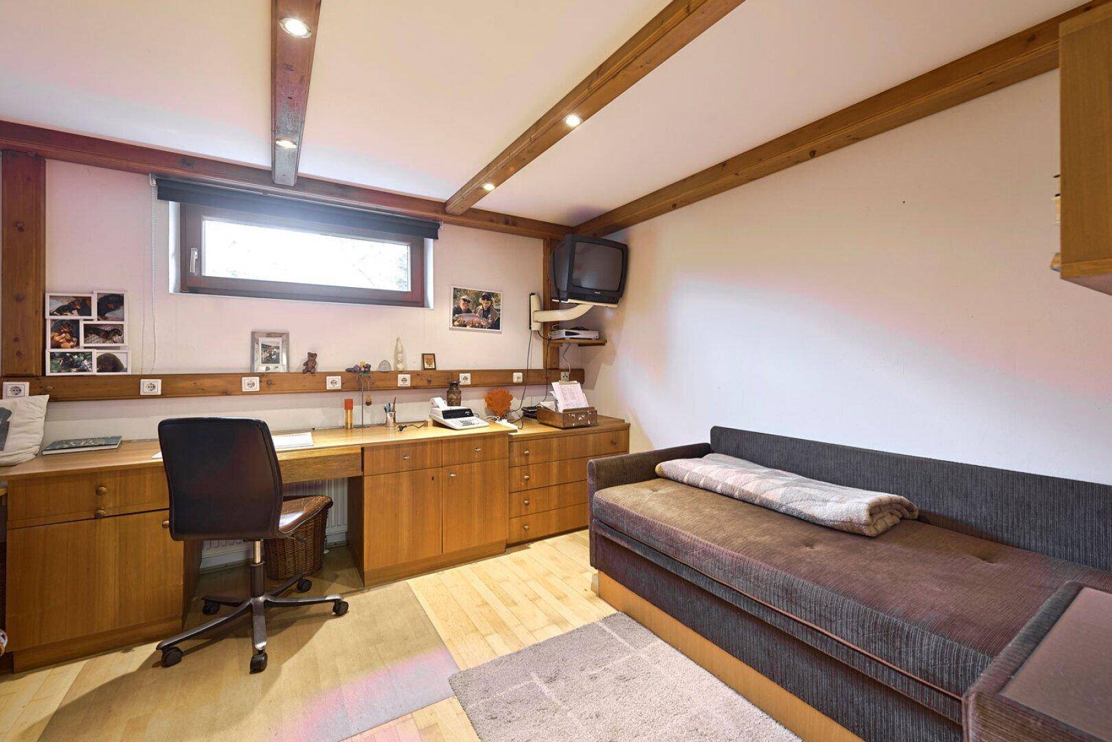 Kellerraum als Büro oder Gästezimmer eingerichtet, mit Holzschreibtisch, Schlafsofa und Holzbalkendecke.