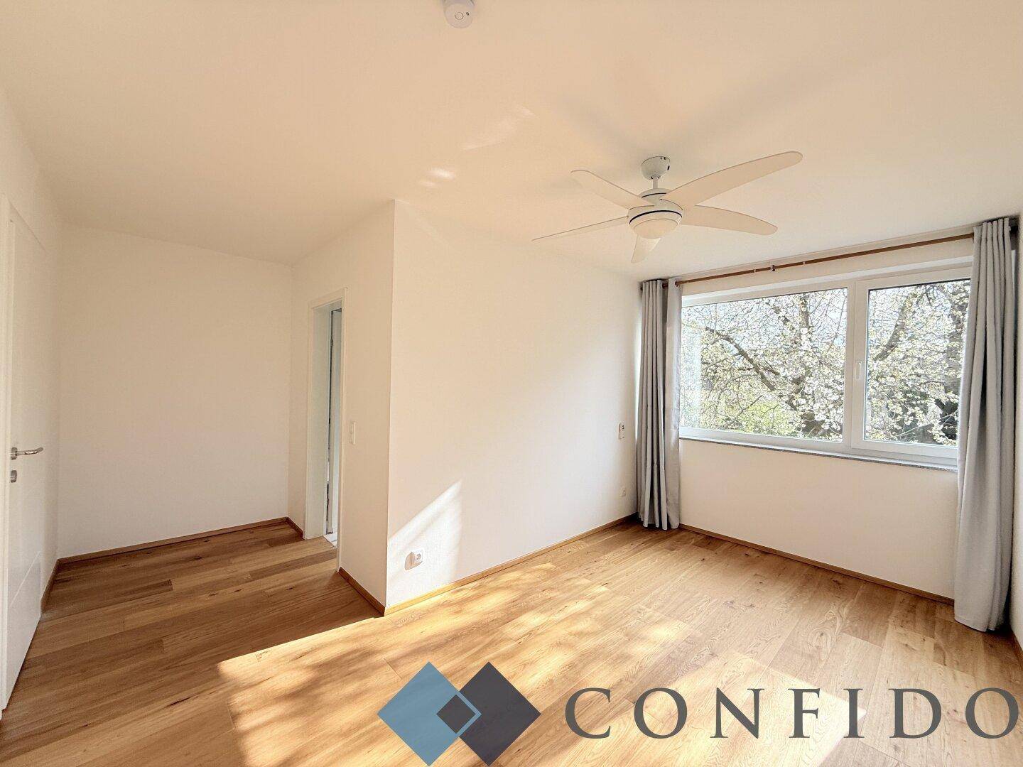 Geräumiges Schlafzimmer mit Holzboden, Deckenventilator und großem Fenster mit Blick ins Grüne.