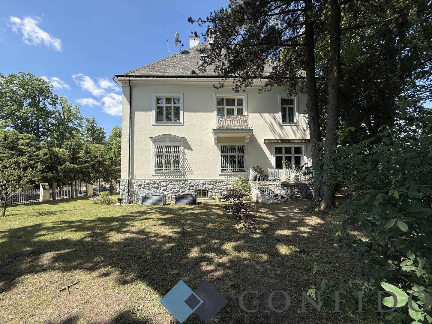 Ansicht der historischen Villa von der Gartenseite mit gepflegtem Rasen und schattenspendenden Bäumen.