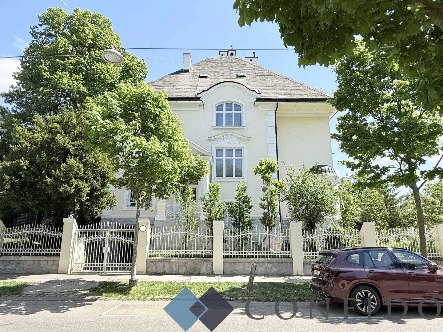 Elegante Straßenansicht der Villa mit schmiedeeisernem Zaun und altem Baumbestand.