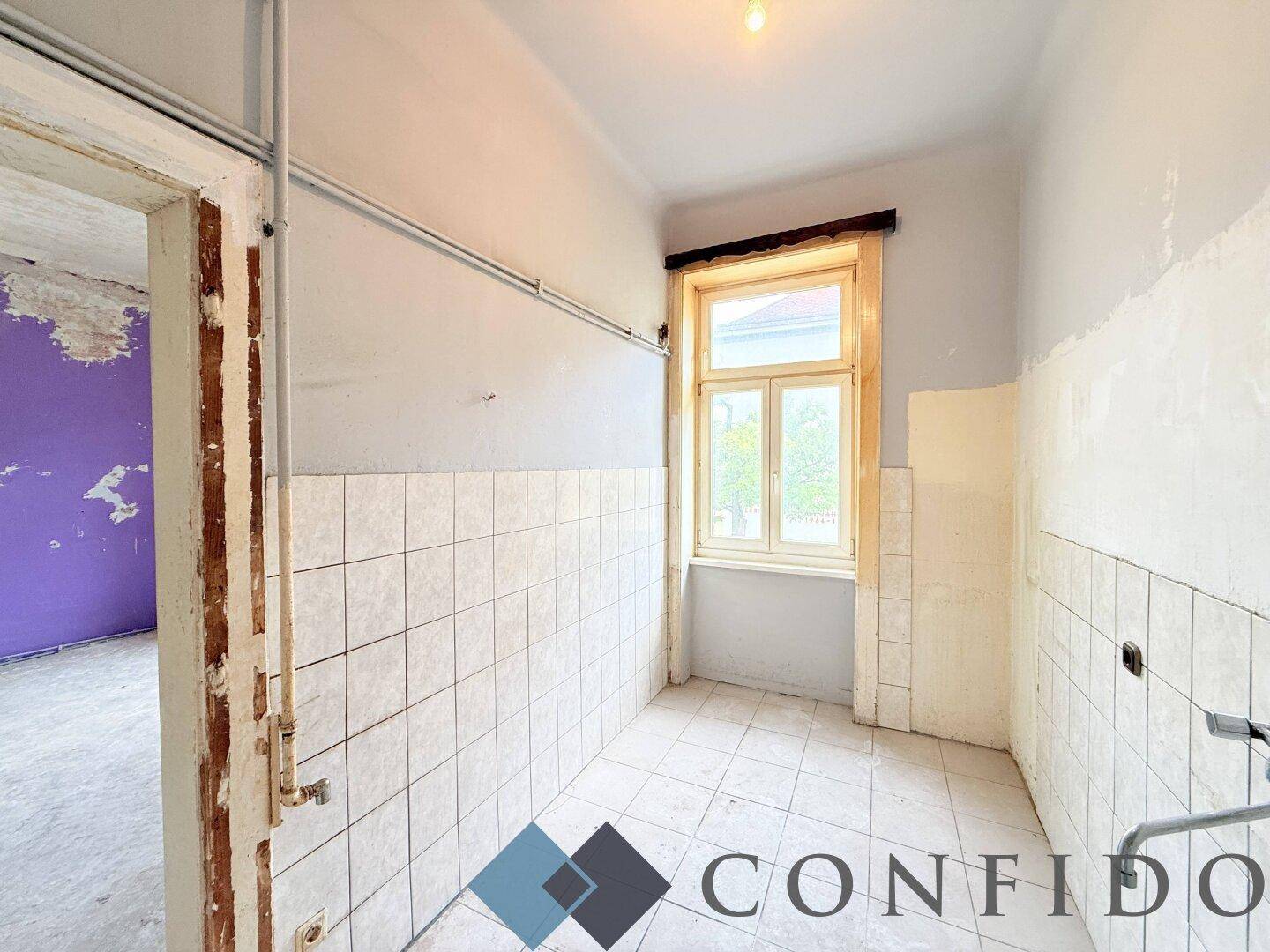 Gefliester Raum mit Fenster und vorbereiteten Anschlüssen, ideal für ein zukünftiges Badezimmer.
