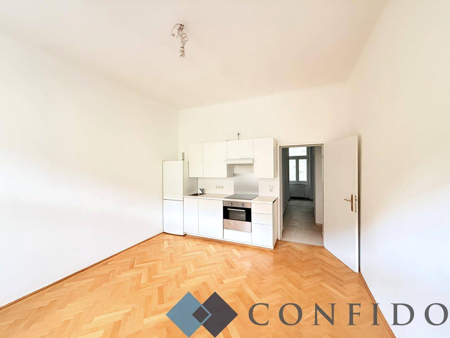 Offener Raum mit integrierter Küchenzeile, Parkettboden und Zugang zu einem weiteren Zimmer.