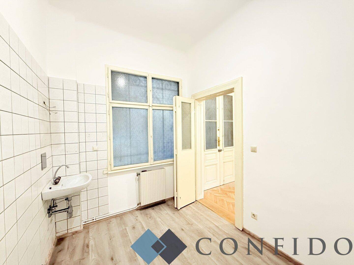 Einfache Küche mit gefliesten Wänden, kleinem Waschbecken und Fenster, bereit für eine Modernisierung.