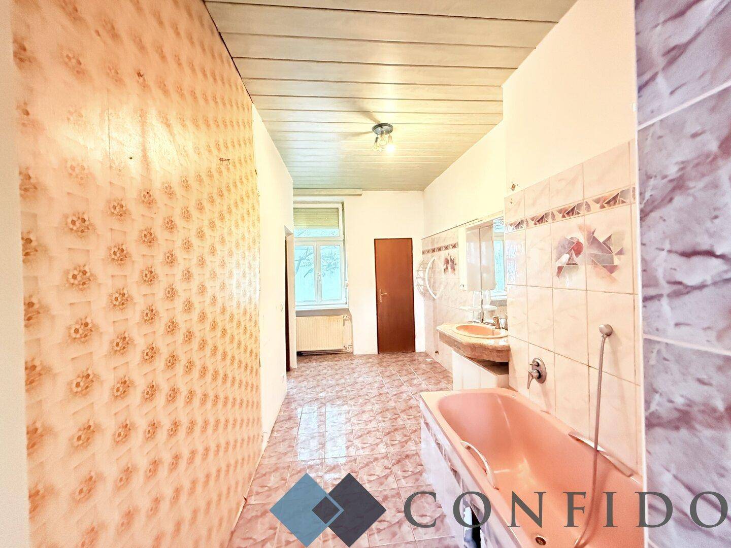 Langes Badezimmer mit rosa Badewanne, Fliesenboden und Fenster am Ende des Raumes.