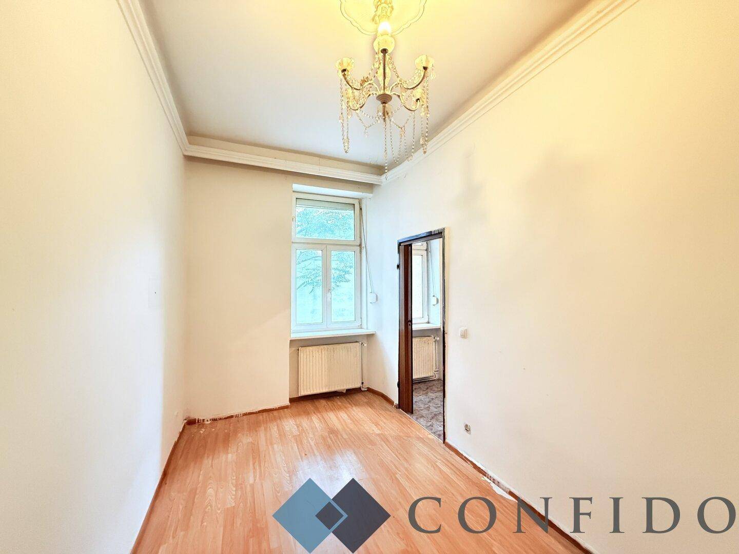 Helles Zimmer mit Parkettboden, Kronleuchter und Fenster, bereit für Renovierung.
